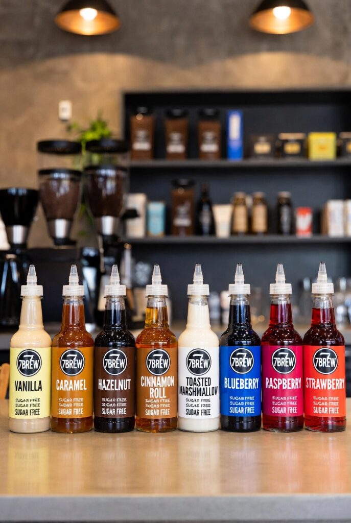 Row of 7 Brew sugar-free syrup bottles including vanilla, caramel, hazelnut, and fruit flavors on counter